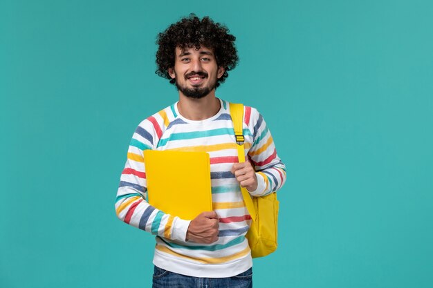 Vista frontal del estudiante masculino con mochila amarilla sosteniendo archivos en la pared azul