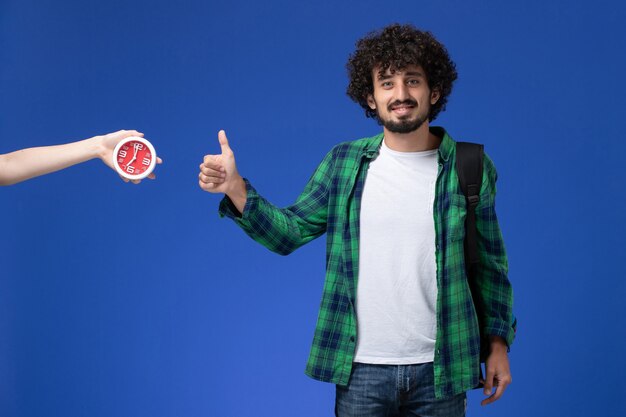 Vista frontal del estudiante masculino con mochila negra sonriendo en la pared azul claro