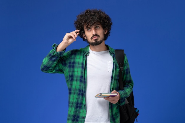 Vista frontal del estudiante masculino con mochila negra sosteniendo el cuaderno y pensando en la pared azul