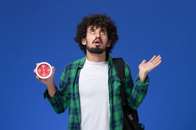 Vista frontal del estudiante masculino con mochila negra sosteniendo relojes en la pared azul claro