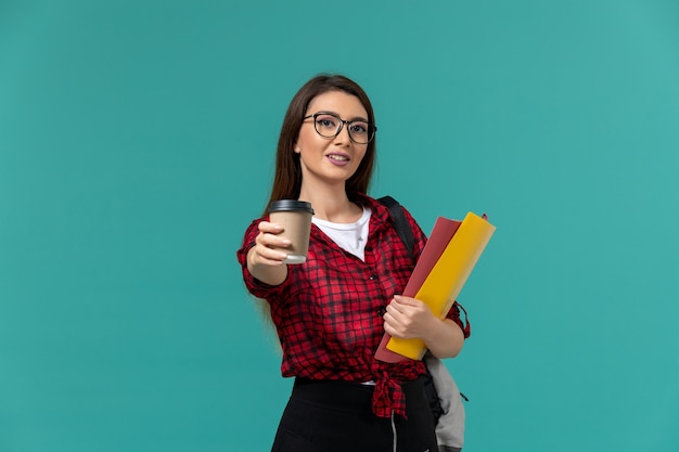 Vista frontal de la estudiante con mochila sosteniendo archivos y café en la pared azul