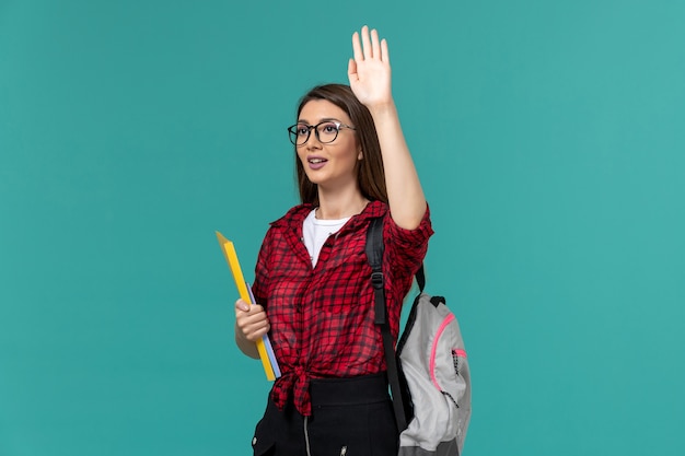 Vista frontal de la estudiante con mochila y sosteniendo archivos levantando su mano sobre la pared azul