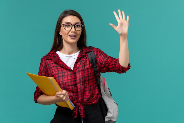 Vista frontal de la estudiante con mochila y sosteniendo archivos en la pared azul claro