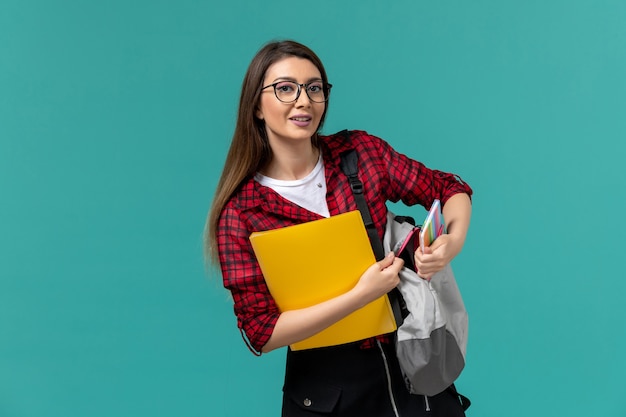 Vista frontal de la estudiante con mochila y sosteniendo archivos en la pared azul claro