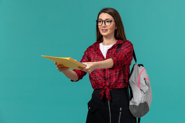 Vista frontal de la estudiante con mochila y sosteniendo archivos en la pared azul claro