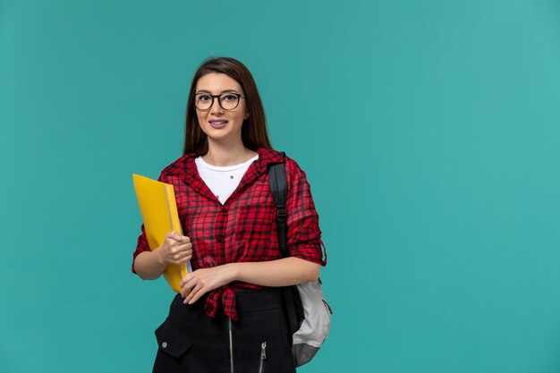 Vista frontal de la estudiante con mochila y sosteniendo archivos en la pared azul