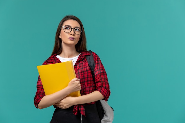 Vista frontal de la estudiante con mochila y sosteniendo archivos pensando en la pared azul claro