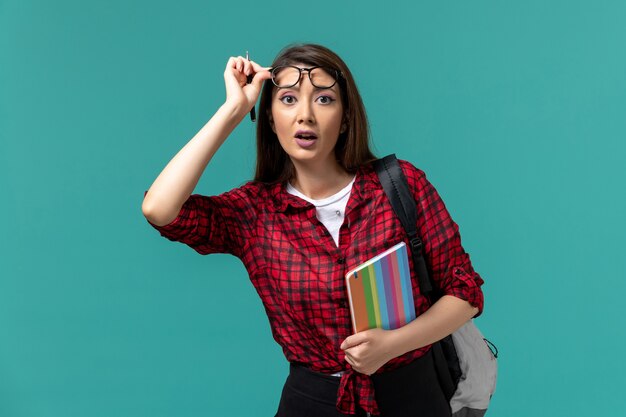 Vista frontal de la estudiante con mochila sosteniendo el cuaderno en la pared azul