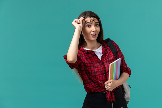 Vista frontal de la estudiante con mochila sosteniendo el cuaderno en la pared azul