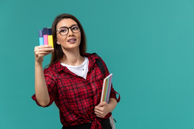 Vista frontal de la estudiante con mochila sosteniendo cuaderno y rotuladores en la pared azul