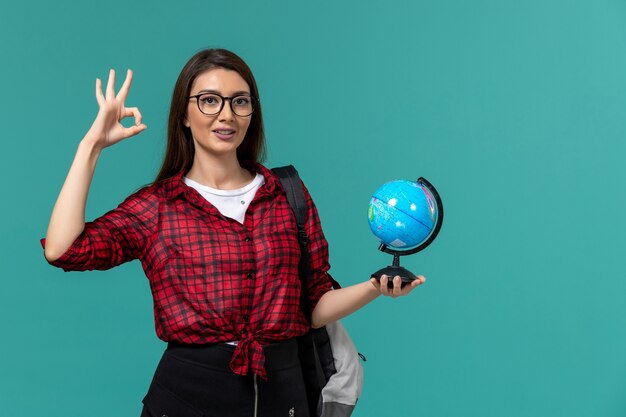 Vista frontal de la estudiante con mochila sosteniendo un pequeño globo en la pared azul claro