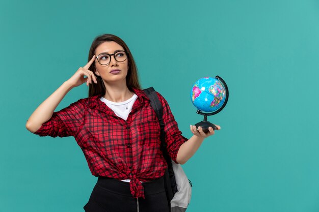 Vista frontal de la estudiante con mochila sosteniendo un pequeño globo pensando en la pared azul claro