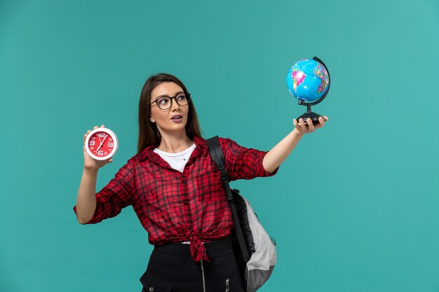 Vista frontal de la estudiante con mochila sosteniendo un pequeño globo y relojes en la pared azul claro