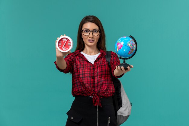 Vista frontal de la estudiante con mochila sosteniendo un pequeño globo y relojes en la pared azul claro