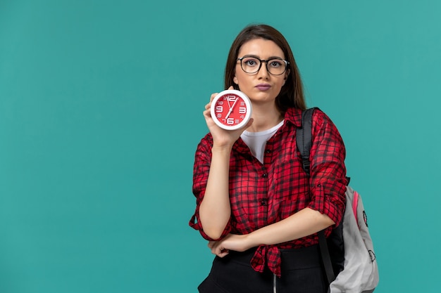 Vista frontal de la estudiante con mochila sosteniendo relojes en la pared azul claro