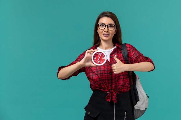 Vista frontal de la estudiante con mochila sosteniendo relojes en la pared azul claro