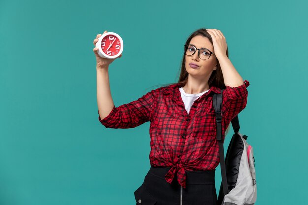 Vista frontal de la estudiante con mochila sosteniendo relojes en la pared azul