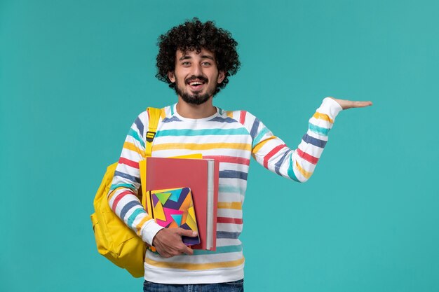 Vista frontal del estudiante varón en camisa a rayas de colores vistiendo mochila amarilla sosteniendo archivos y cuadernos en la pared azul
