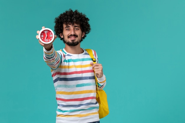 Vista frontal del estudiante varón con mochila amarilla sosteniendo relojes en la pared azul claro