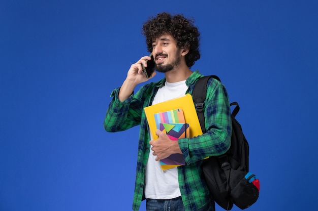 Vista frontal del estudiante varón con mochila negra sosteniendo cuadernos y archivos hablando por teléfono en la pared azul claro