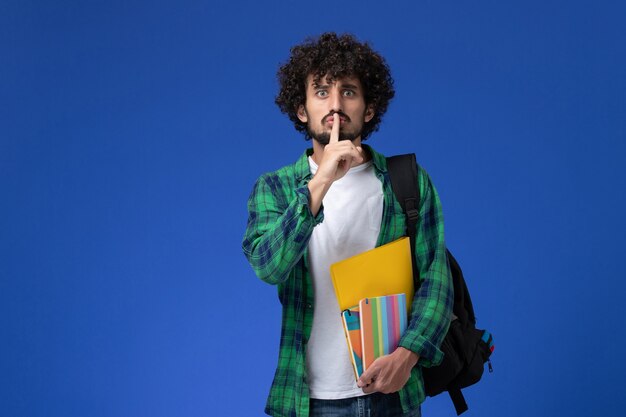 Vista frontal del estudiante varón con mochila negra sosteniendo cuadernos y archivos en la pared azul