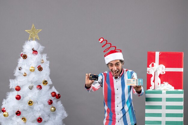 Vista frontal eufórico joven sosteniendo una tarjeta y un boleto de viaje alrededor del árbol de Navidad y regalos