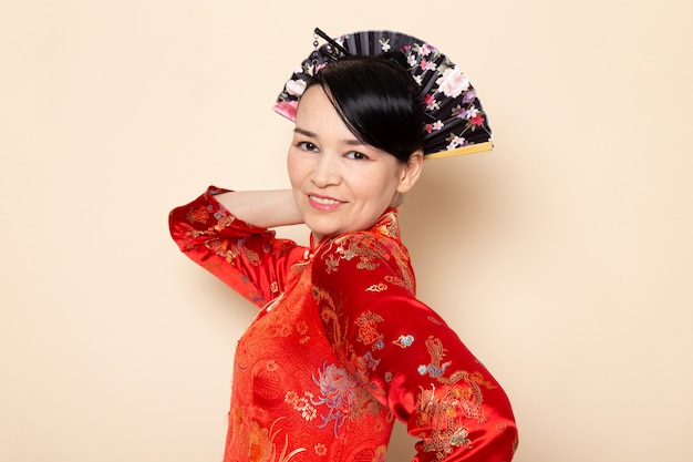 Una vista frontal exquisita geisha japonesa en el tradicional vestido rojo japonés con palos para el pelo posando sosteniendo abanico plegable elegante sonriendo en la ceremonia de fondo crema Japón