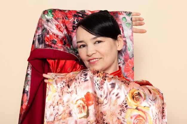 Foto gratuita una vista frontal exquisita geisha japonesa en vestido tradicional rojo japonés posando con tejido de flores diseñado elegante sonriendo en la ceremonia de fondo crema japón