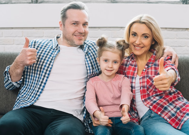 Vista frontal de la familia feliz que muestra el pulgar hacia arriba signo sentado en el sofá