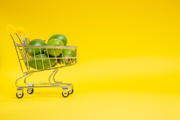 Vista frontal de la feijoa verde fresca dentro de la pequeña canasta móvil en la superficie amarilla de frutas exóticas suaves de color