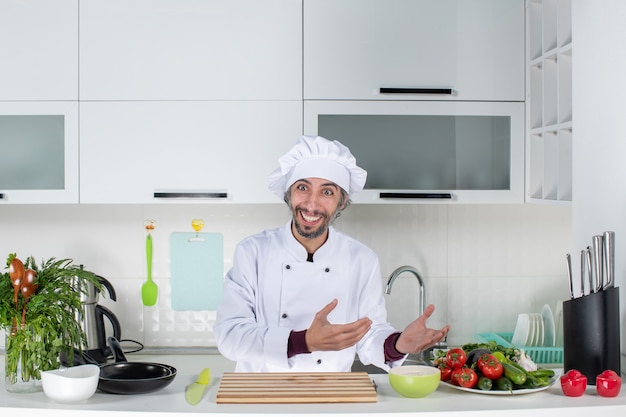 Vista frontal feliz chef masculino en uniforme de pie detrás de la mesa de la cocina