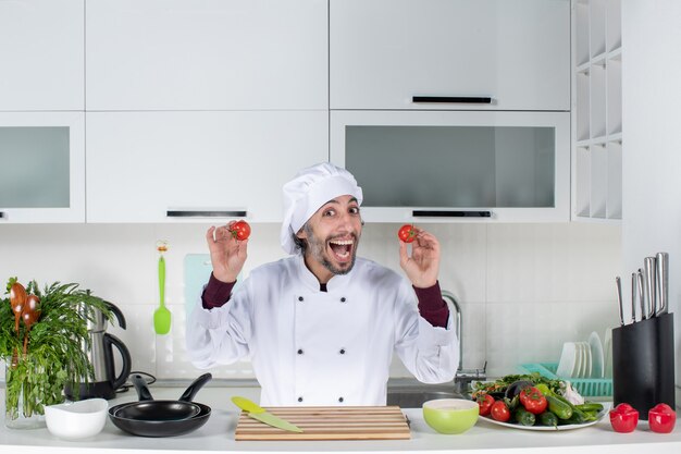 Vista frontal feliz chef masculino en uniforme sosteniendo tomates en la cocina
