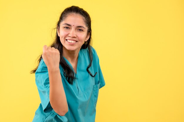 Vista frontal feliz doctora en uniforme mostrando gesto ganador sobre fondo amarillo aislado