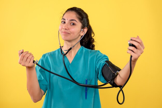 Vista frontal feliz doctora en uniforme sosteniendo esfigmomanómetros de pie sobre fondo amarillo