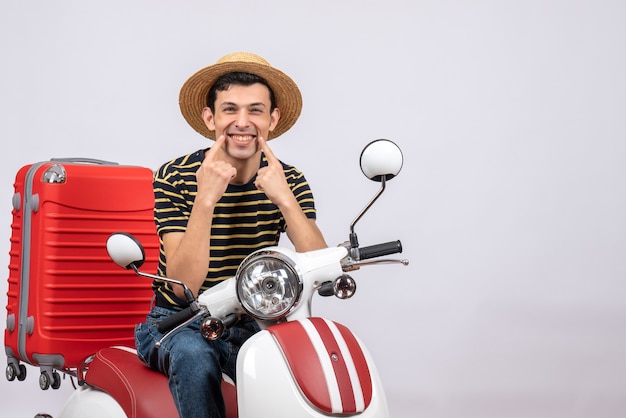 Vista frontal feliz joven con sombrero de paja en ciclomotor apuntando a su sonrisa