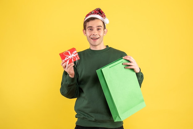 Vista frontal feliz joven con sombrero de santa con bolsa de compras verde y pie de regalo en amarillo