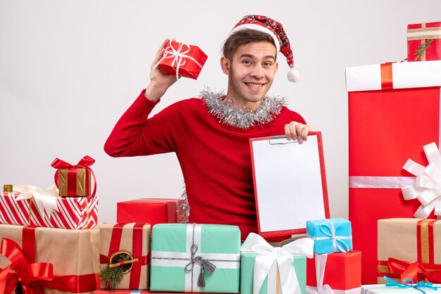 Vista frontal feliz joven sosteniendo portapapeles sentado alrededor de regalos de Navidad