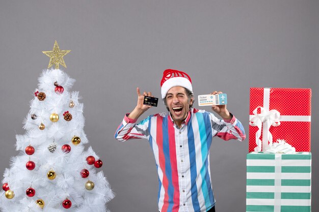 Vista frontal feliz joven sosteniendo la tarjeta y el boleto de viaje alrededor del árbol de Navidad