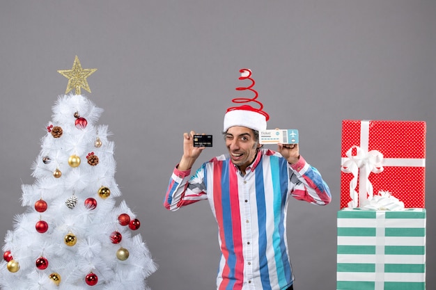 Vista frontal feliz joven con tarjeta de tenencia de ojos parpadeados y boleto de viaje alrededor del árbol de Navidad y regalos