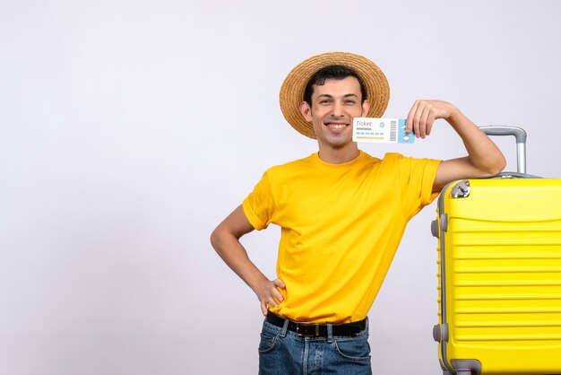 Vista frontal feliz joven turista de pie cerca de la maleta amarilla poniendo la mano en la cintura sosteniendo un boleto aéreo