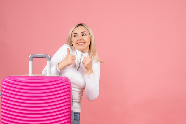 Vista frontal feliz mujer rubia con maleta rosa dando pulgar hacia arriba