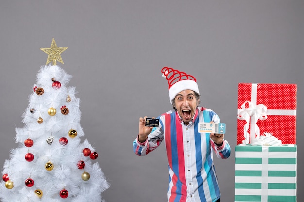 Vista frontal feliz Navidad hombre con tarjeta y billete de viaje alrededor del árbol de Navidad y regalos