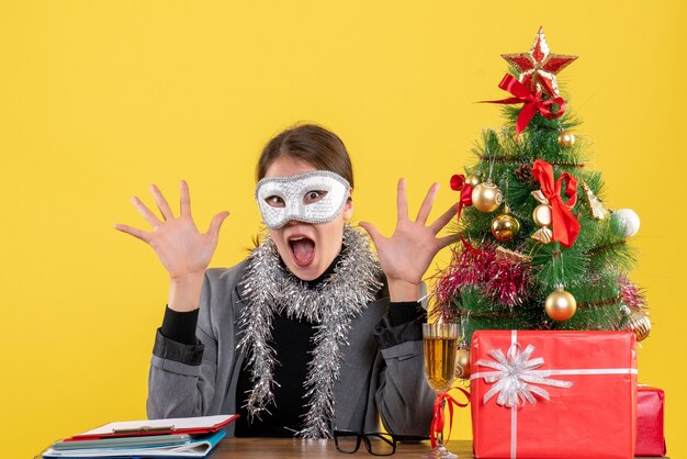 Vista frontal feliz niña con máscara sentada en la mesa abriendo ambas manos árbol de Navidad y cóctel de regalos