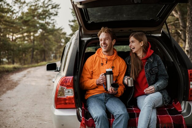 Vista frontal de la feliz pareja disfrutando de una bebida caliente en el maletero del coche