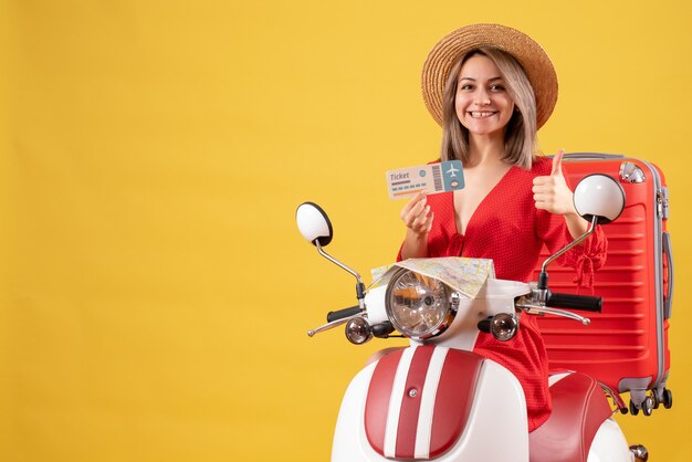 Vista frontal de la feliz señorita en vestido rojo con boleto dando pulgar hacia arriba en ciclomotor