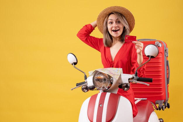 Vista frontal de la feliz señorita en vestido rojo con tarjeta bancaria en ciclomotor