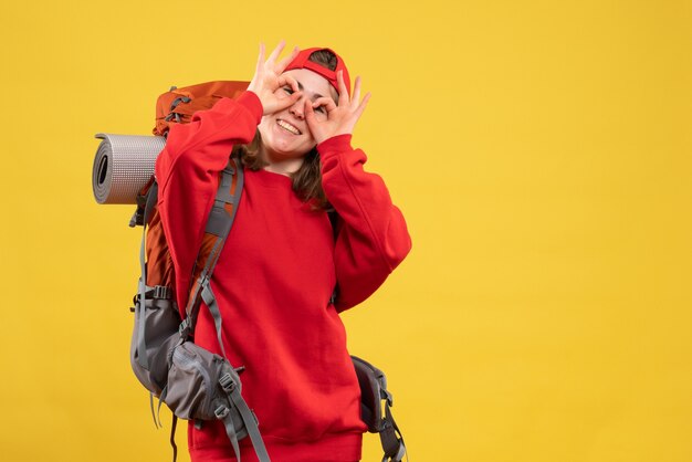 Vista frontal feliz viajera con mochila poniendo signo de ok delante de sus ojos