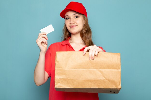 Una vista frontal femenina atractiva mensajero en polo rojo gorra roja y jeans con paquete y tarjeta blanca posando sonriente sobre el fondo azul trabajo de servicio de alimentos