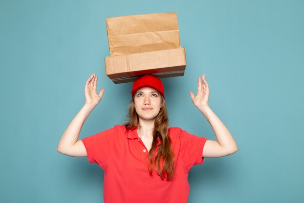 Una vista frontal femenina atractiva mensajero en polo rojo gorra roja con paquetes marrones por encima de su cabeza posando en el trabajo de servicio de alimentos de fondo azul