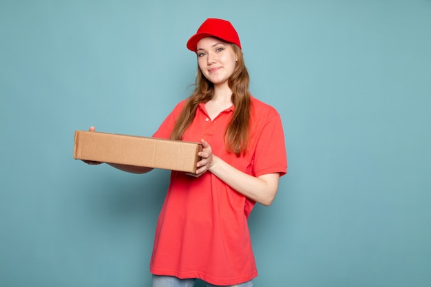 Una vista frontal femenino atractivo mensajero en polo rojo gorra roja y jeans con paquete posando en el trabajo de servicio de alimentos de fondo azul